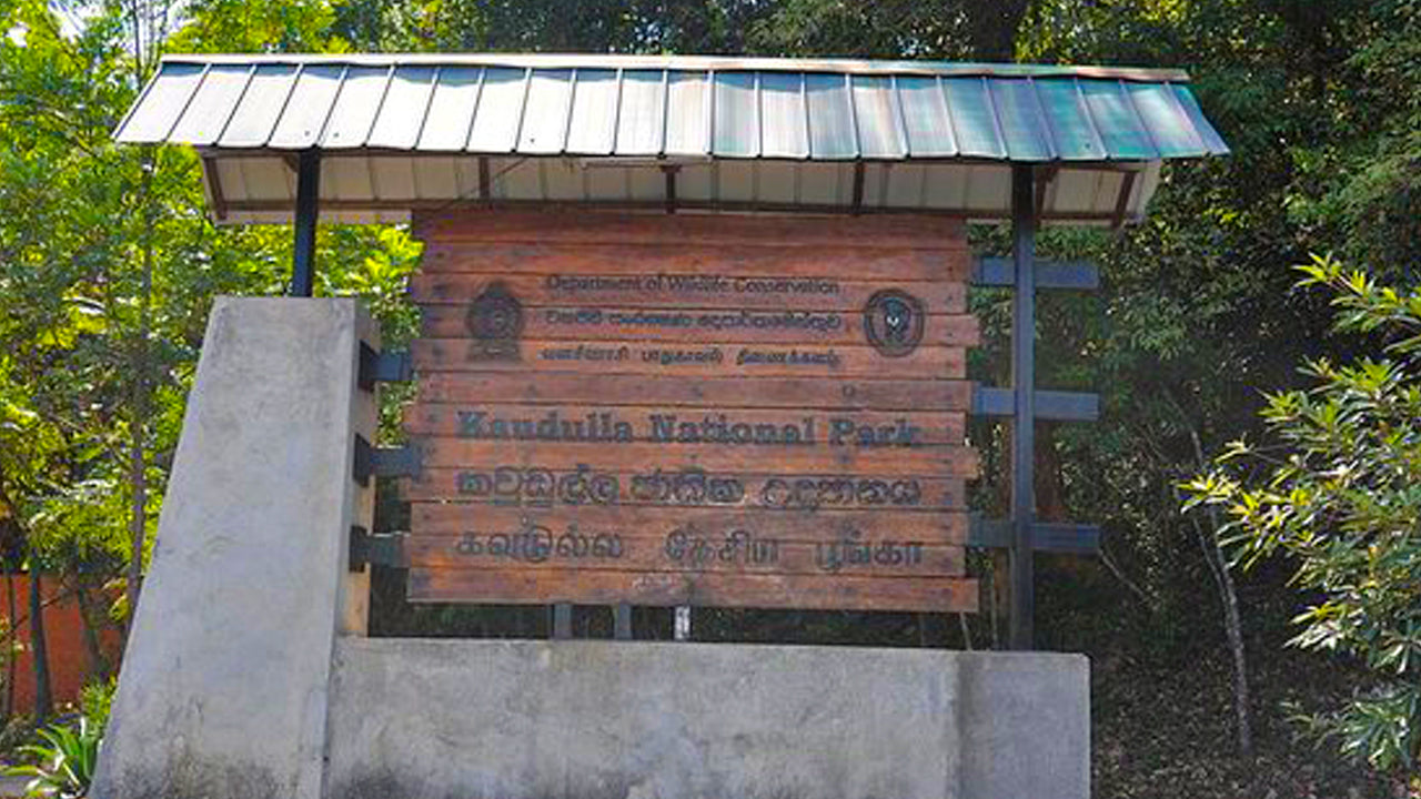 Entrance gate to Kaudulla National Park with a wooden signboard providing information in English and Sinhala.